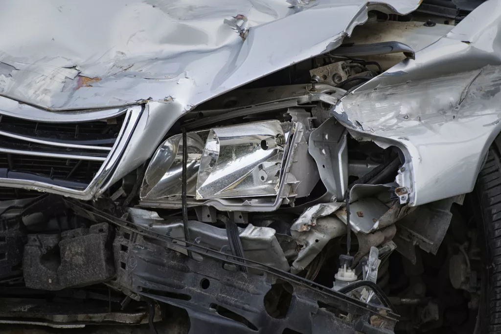 An damaged silver sedan with significant front-end destruction, including a crushed bumper and exposed engine parts, following a car accident.