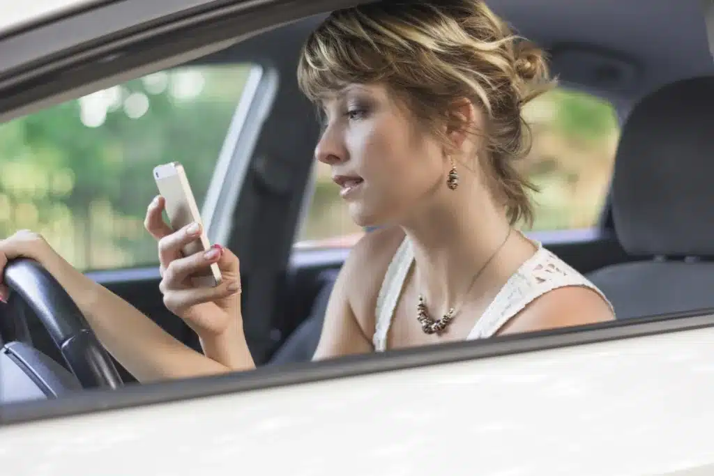 A blonde woman looking down at her smartphone while holding the steering wheel of a car, illustrating the dangers of distracted driving accidents.