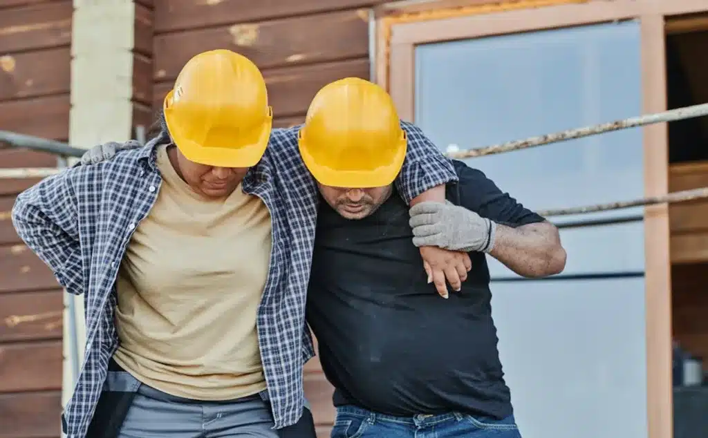 A male worker wearing a plaid shirt and yellow hard hat helps an injured coworker in a black t-shirt and yellow hard hat on a construction site, illustrating the risks in the most dangerous US cities to work in.