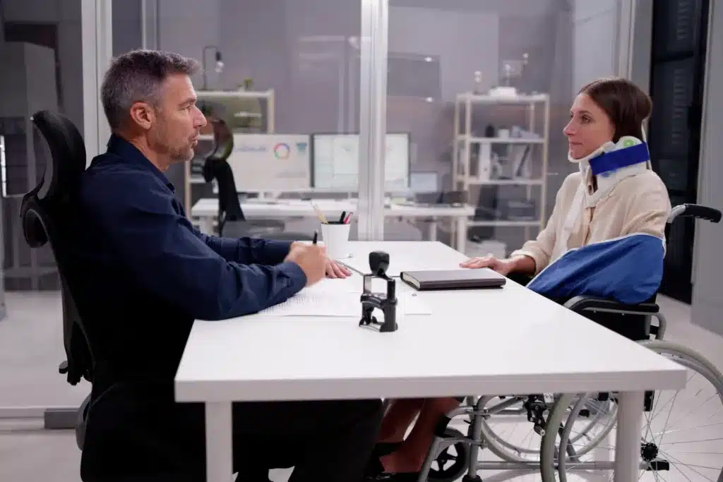 A male attorney or insurance adjuster sits at a desk discussing a workers' compensation claim with an injured female worker wearing a neck brace and arm sling.
