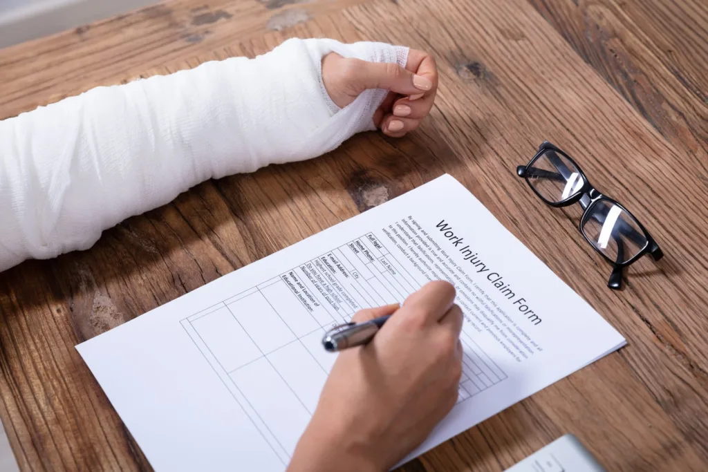 A person with an arm cast filling out a work injury claim form next to eyeglasses on a wooden table.