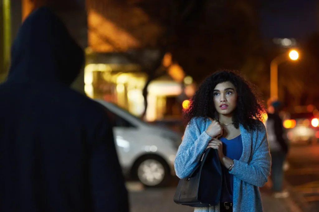 A worried young woman clutching her bag on a dark street while a shadowy figure approaches, representing negligent security leading to sexual assault.