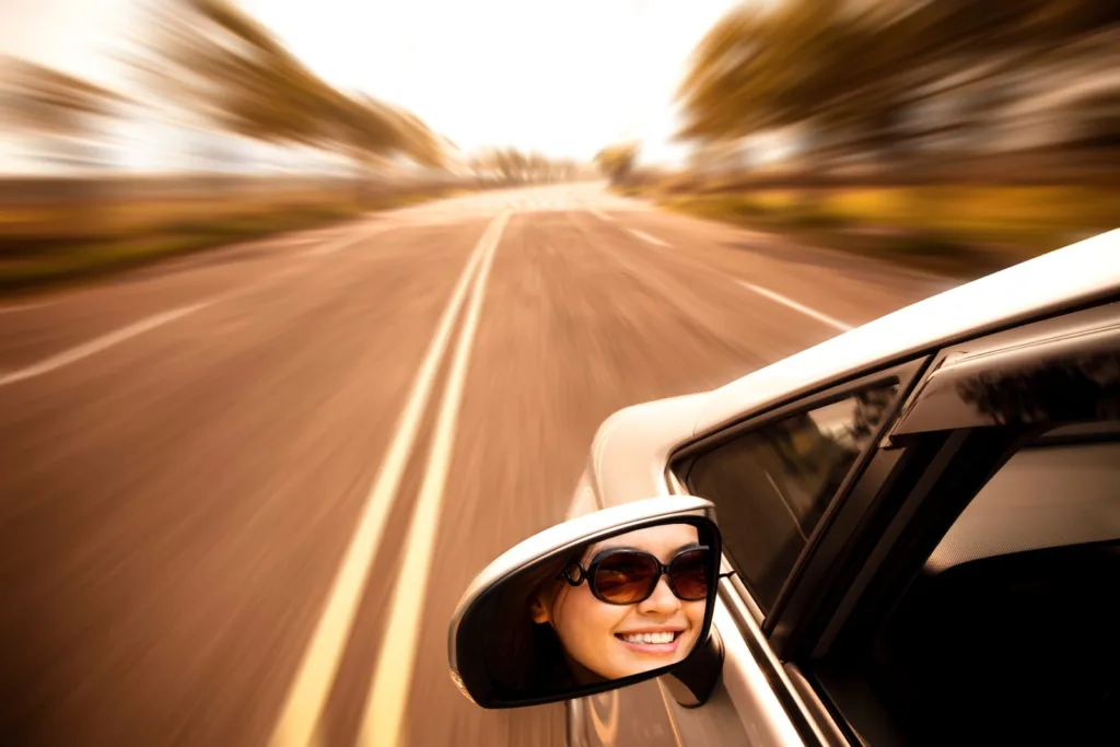 A close-up shot of a car side mirror reflecting a smiling woman wearing sunglasses as she enjoys a drive on an open road, highlighting the importance of personal injury protection and auto insurance for Texas drivers.